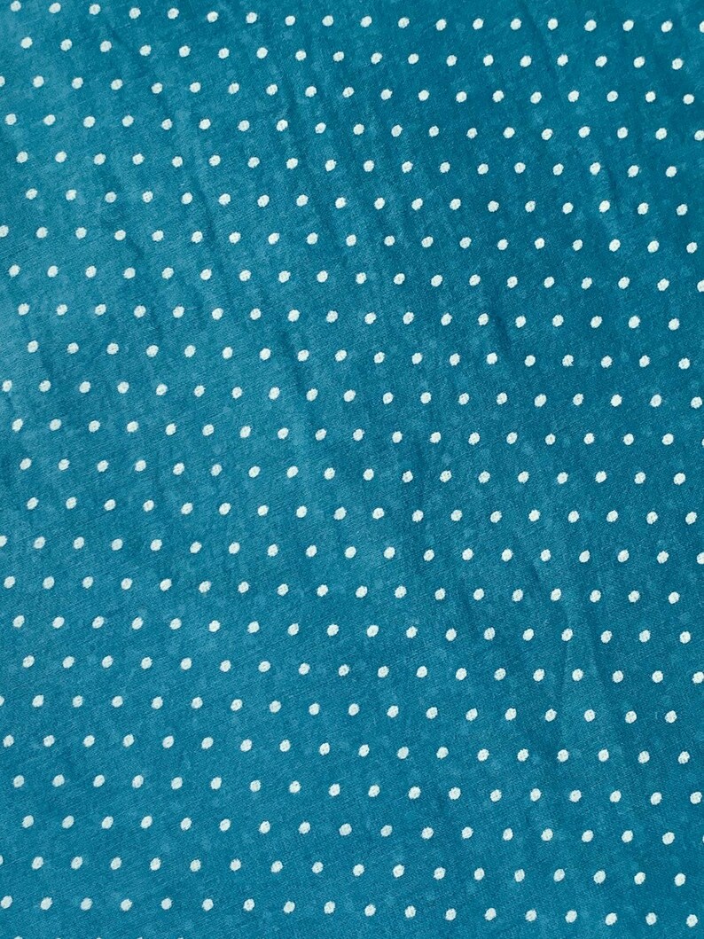 May include: A teal fabric featuring a repeating pattern of small, white, evenly spaced polka dots. The material has a slightly textured surface with a consistent weave, displaying a uniform distribution of the white dots across the blue background.