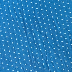 May include: A bright blue fabric featuring a repeating pattern of small, white, circular polka dots distributed evenly across the entire surface. The material displays a soft, slightly textured appearance with subtle folds and creases throughout.