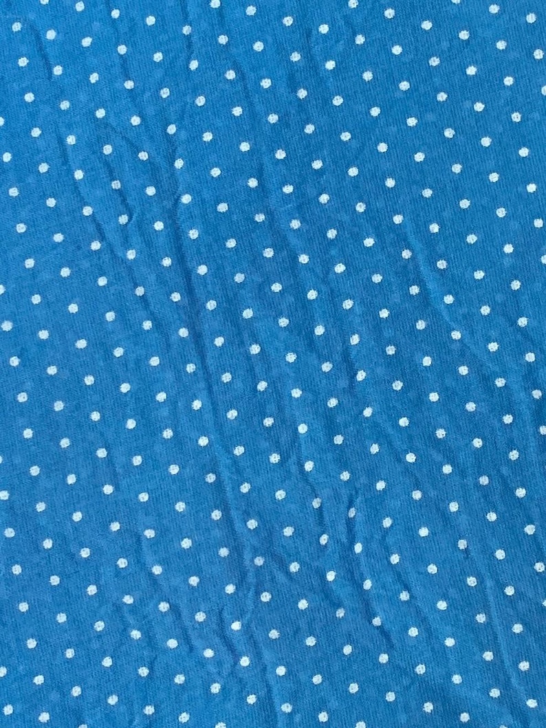 May include: A bright blue fabric featuring a repeating pattern of small, white, circular polka dots distributed evenly across the entire surface. The material displays a soft, slightly textured appearance with subtle folds and creases throughout.
