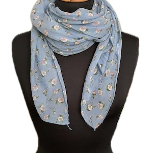 May include: A light blue scarf with a delicate floral pattern of small pink and white roses. The scarf is draped over a black mannequin, showcasing its flowing fabric and design. A stylish accessory for any outfit.