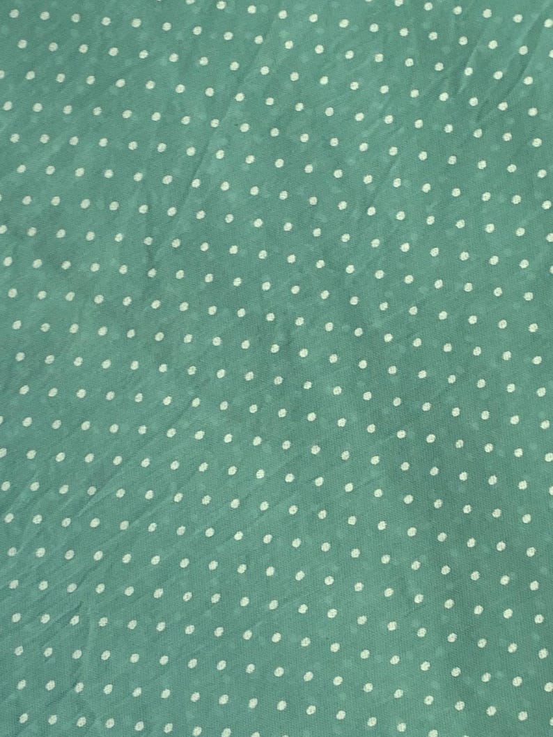 May include: A light green fabric featuring a repeating pattern of small, white, circular polka dots distributed evenly across the surface. The material has a slightly textured, semi-sheer appearance with subtle folds and creases throughout the textile.