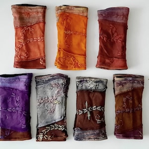 Op de afbeelding: Acht kleurrijke armwarmers in tinten oranje, rood, paars en grijs worden getoond tegen een witte achtergrond. Elke armwarmer heeft een patchwork ontwerp met geborduurde bloempatronen. De armwarmers zijn gemaakt van een zachte, getextureerde stof.