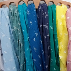 May include: A collection of colorful scarves, each displaying a dragonfly pattern. The scarves are arranged in a rainbow of hues, including white, blue, green, yellow, pink, and red. The dragonfly design is a delicate, repeating pattern in white.