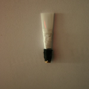 Puede incluir: Una caña de clarinete blanca con una línea roja en el centro. La caña está envuelta en cinta blanca y tiene una base negra y dorada.