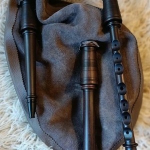 Può includere: Un set di cornamuse in legno marrone scuro con una sacca in morbida pelle scamosciata grigia. Lo strumento include un chanter con fori per le dita e due bordoni, tutti realizzati in legno scuro liscio con anelli torniti dettagliati.