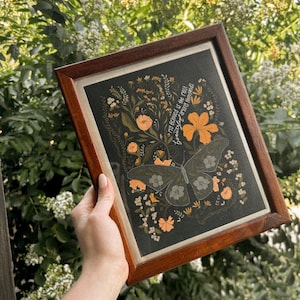 May include: A rectangular wooden frame containing a black print featuring a large butterfly with patterned wings surrounded by orange and white wildflowers and foliage. The text "My Beloved is the most beautiful among thousands" is printed in white script.