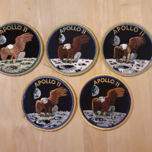 May include: Five round embroidered patches with a dark blue background, a white moon, and a brown eagle with outstretched wings. Each patch has a gold border and the text "APOLLO 11" in white.