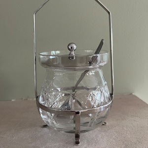 May include: A clear glass jar with a cut crystal design, a silver lid, and a silver spoon. The jar is sitting on a silver stand with a handle.