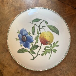 May include: A white ceramic plate with a green and red border. The plate features a hand-painted design of a blue flower, green leaves, and a yellow and red apple.
