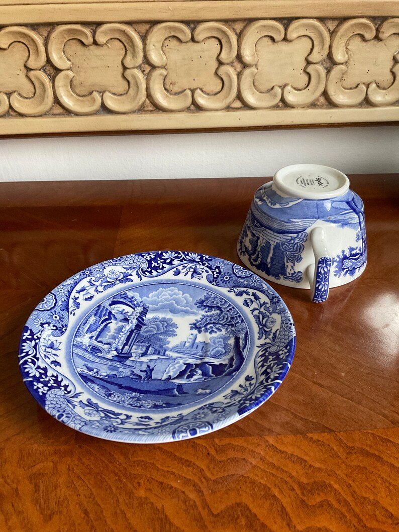 May include: Blue and white porcelain teacup and saucer set with a floral pattern. The saucer features a landscape scene with a building and trees. The teacup has a matching design and a white handle.