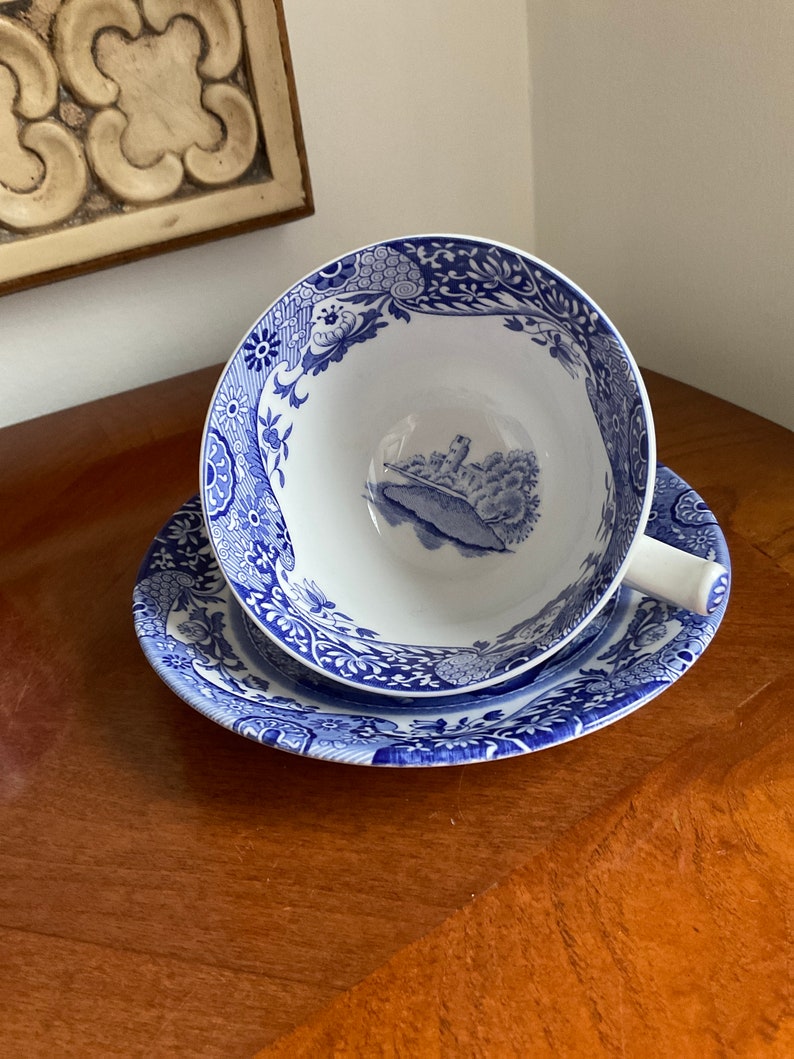 May include: A blue and white teacup and saucer set with a floral pattern. The saucer has a scalloped edge. The teacup has a handle and a scene of a building and trees in the centre.