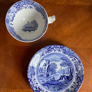 May include: A blue and white porcelain teacup and saucer set with a floral pattern and a landscape scene. The saucer features a detailed depiction of a bridge, trees, and a building.