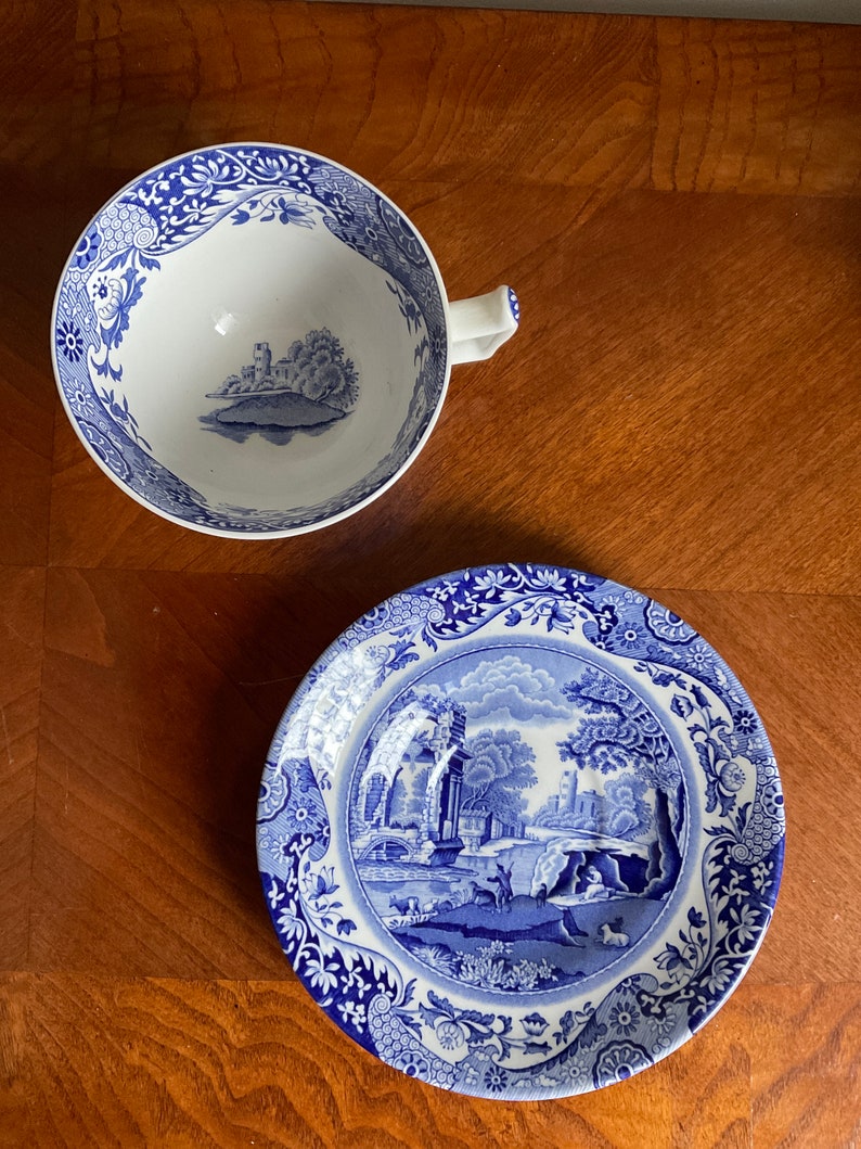 May include: A blue and white porcelain teacup and saucer set with a floral pattern and a landscape scene. The saucer features a detailed depiction of a bridge, trees, and a building.