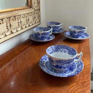May include: Four blue and white teacups and saucers with a floral pattern. The cups and saucers are arranged on a wooden table.