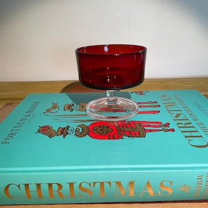 May include: A ruby red glass dessert dish with a clear glass stem and base. The dish is sitting on top of a teal blue book titled "Christmas & Winter Tales" by Peter Wentzel.