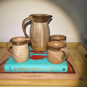 May include: Set of four brown and white ceramic mugs with a brown and white ceramic jug. The mugs and jug have a rustic, handmade look. The set is displayed on a wooden table with a book titled "Christmas" underneath.