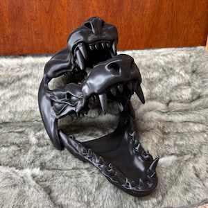 May include: A black, 3D-printed alligator skull mask with detailed teeth and a textured surface. The mask features an open mouth and is designed to be worn. The mask is displayed on a gray, furry surface.