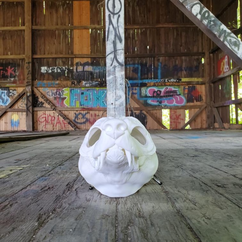 May include: A white, detailed skull sculpture with large eye sockets and sharp teeth. The skull is positioned on a weathered wooden surface, with a graffiti-covered wooden structure in the background. The sculpture is a unique decorative item.