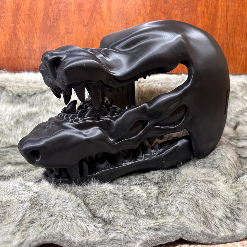 May include: A black, 3D-printed sculpture of a stylized animal skull, featuring sharp teeth and detailed bone structure. The sculpture is displayed on a textured, gray and white surface, with a wooden background.