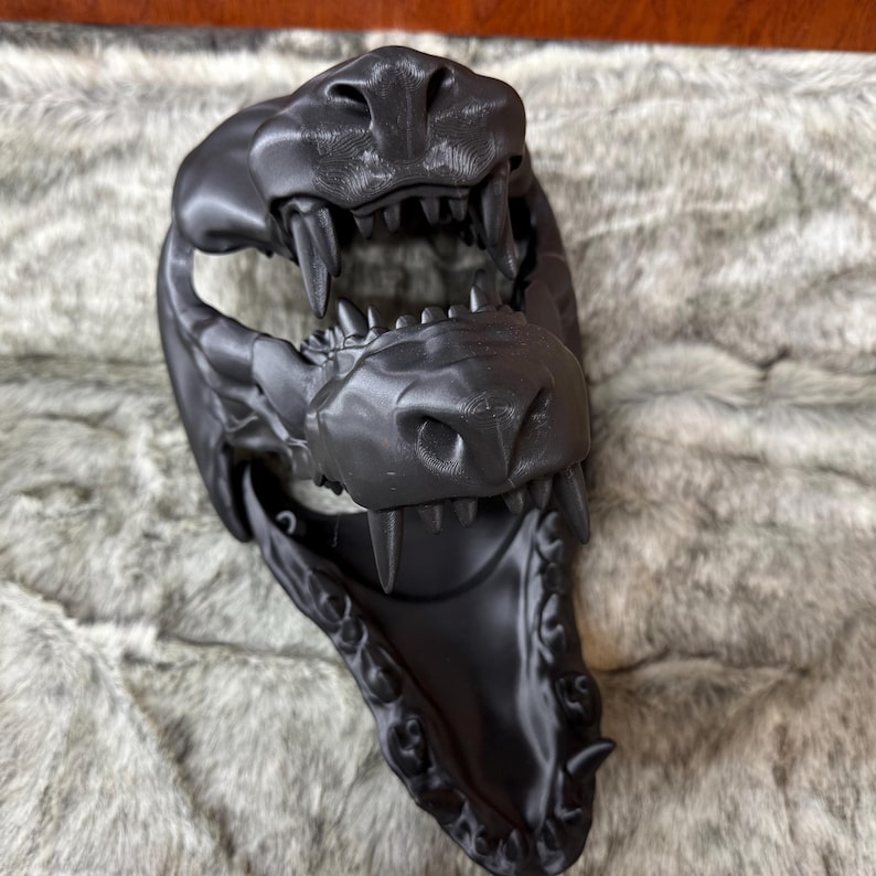 May include: A black, 3D-printed mask depicting a stylized animal skull with sharp teeth and detailed facial features. The mask is designed to be worn, with an open mouth and eye sockets. The mask is made of a matte black material.