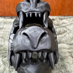 May include: A black, 3D-printed dragon skull with detailed teeth and textured surfaces. The skull is displayed against a backdrop of faux fur and a wooden surface. The dragon skull is a decorative item.