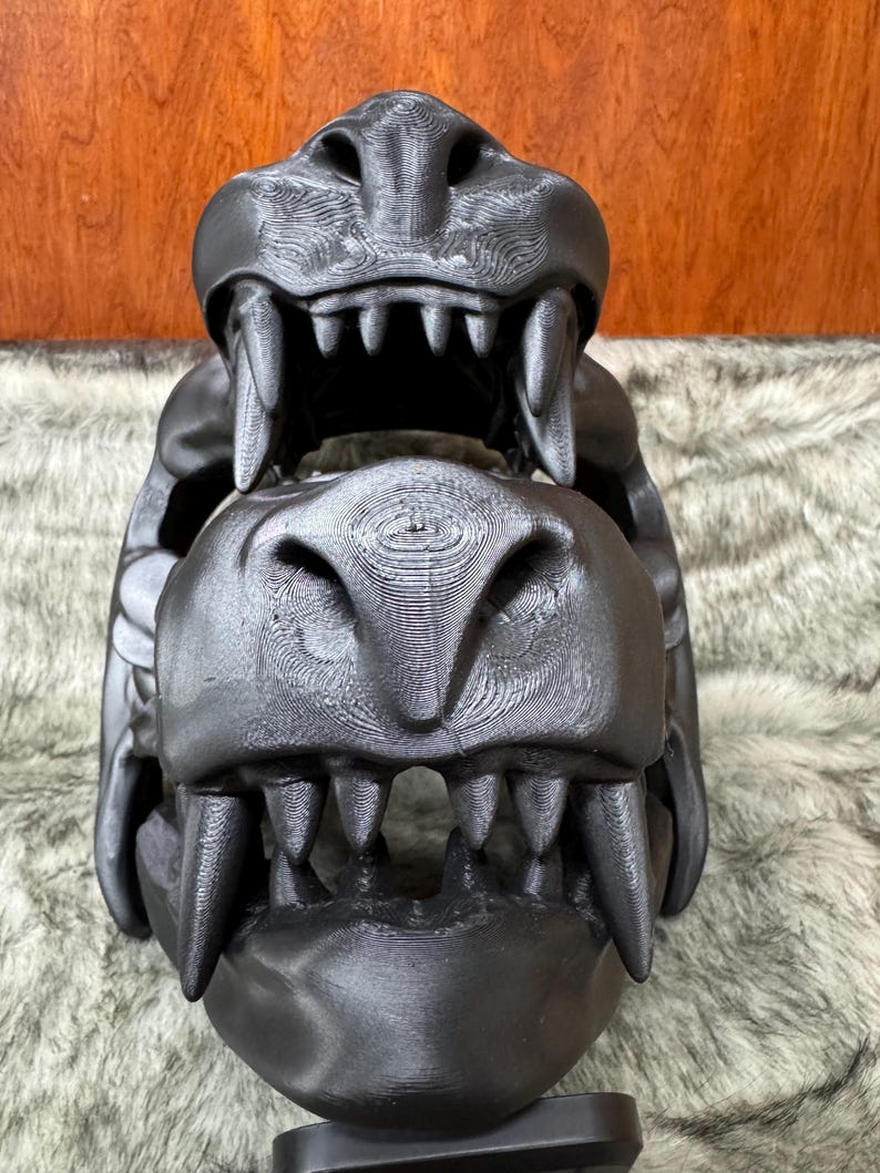 May include: A black, 3D-printed dragon skull with detailed teeth and textured surfaces. The skull is displayed against a backdrop of faux fur and a wooden surface. The dragon skull is a decorative item.