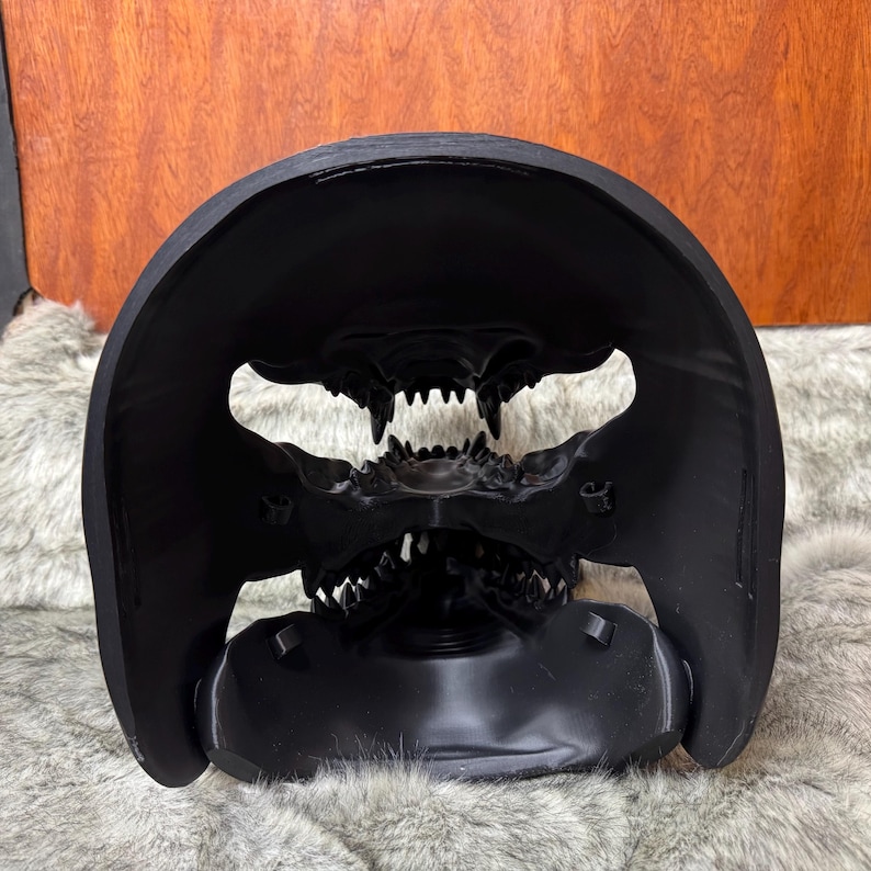 May include: A black, stylized skull mask with sharp teeth and a wide, open mouth. The mask has a curved, protective top and is set against a textured, neutral background. The design is detailed and menacing.