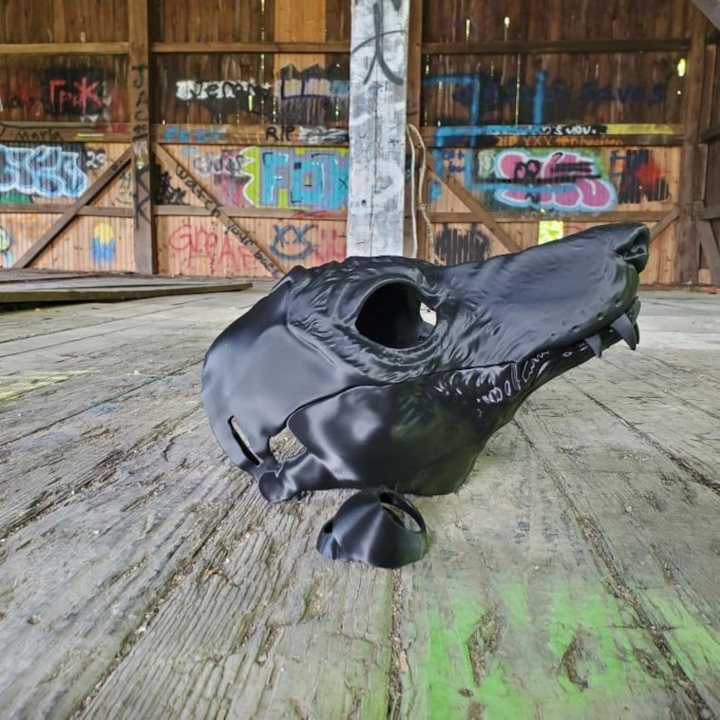 May include: A black, 3D-printed wolf skull mask with detailed features, including eye sockets and teeth. The mask is displayed on a weathered wooden surface, with a detached lower jaw piece nearby. The background shows a graffiti-covered wooden structure.