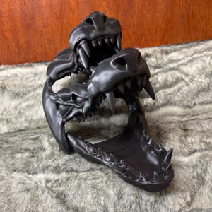 May include: A black, 3D-printed dragon skull sculpture with sharp teeth and detailed bone structure. The sculpture features two dragon heads, one above the other, with open jaws. The piece is displayed on a textured, gray surface.