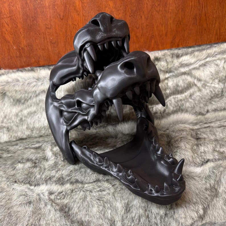 May include: A black, 3D-printed dragon skull sculpture with sharp teeth and detailed bone structure. The sculpture features two dragon heads, one above the other, with open jaws. The piece is displayed on a textured, gray surface.