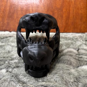 May include: A black, 3D-printed animal skull with open jaws and visible teeth. The skull is resting on a gray, faux fur surface. The skull's details are clearly defined, showcasing the texture and form of the design.
