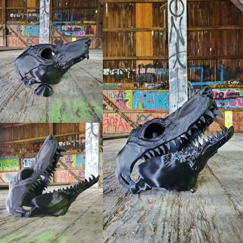 May include: Black 3D-printed dinosaur skull mask with sharp teeth and eye sockets. The mask is designed to be worn and has a detailed, textured surface. The mask is displayed in a rustic, graffiti-covered environment.
