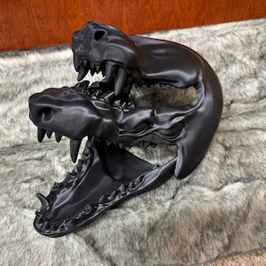 May include: A black, 3D-printed sculpture of two animal skulls, one atop the other, with open jaws and visible teeth. The sculpture is detailed and sits on a faux fur surface.