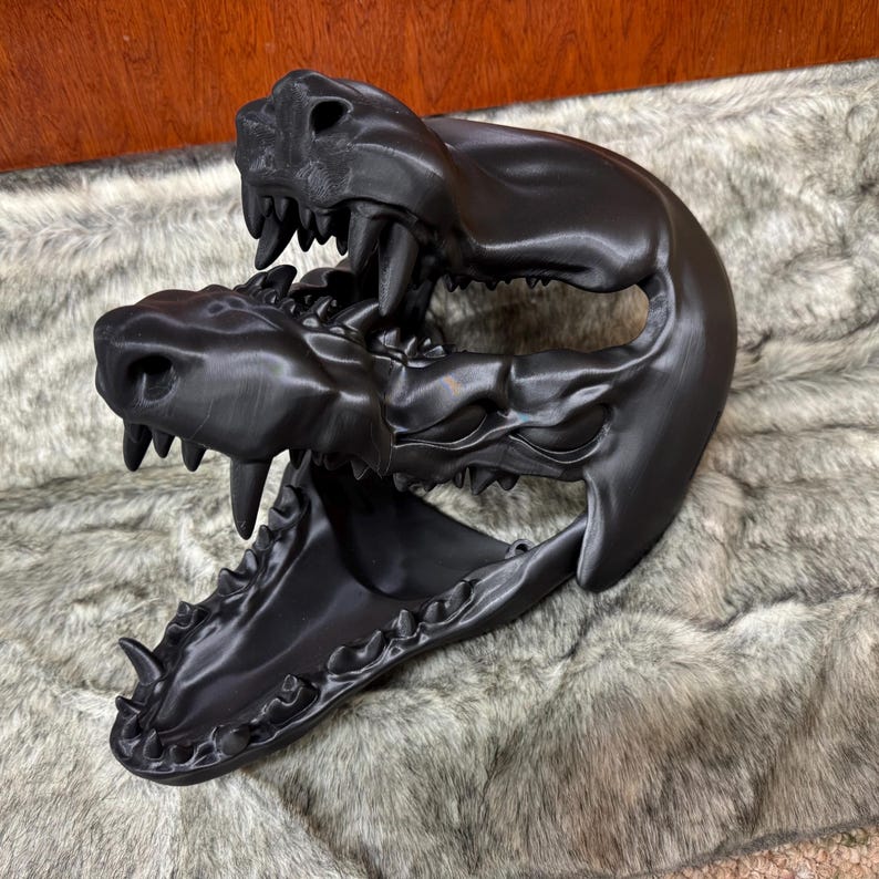 May include: A black, 3D-printed sculpture of two animal skulls, one atop the other, with open jaws and visible teeth. The sculpture is detailed and sits on a faux fur surface.