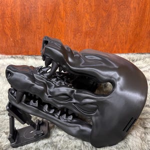 May include: A black, stylized mask shaped like a snarling beast's head, featuring sharp teeth and detailed facial features. The mask is supported by a black stand, and rests on a furry surface.