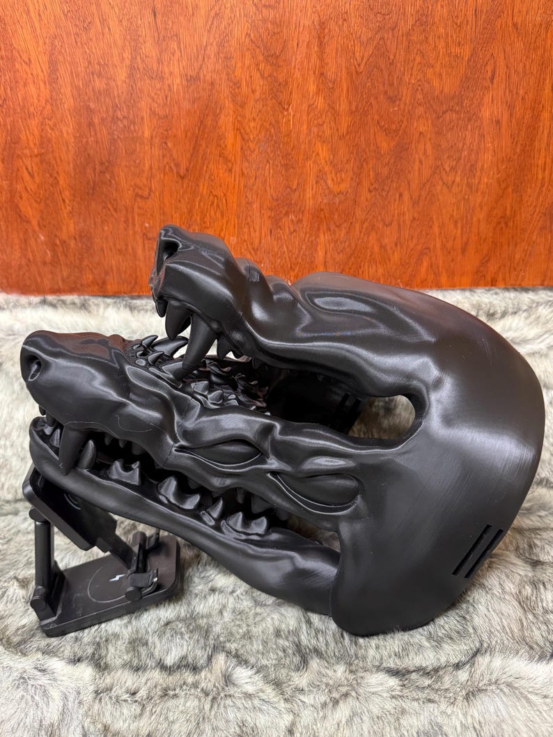 May include: A black, stylized mask shaped like a snarling beast's head, featuring sharp teeth and detailed facial features. The mask is supported by a black stand, and rests on a furry surface.
