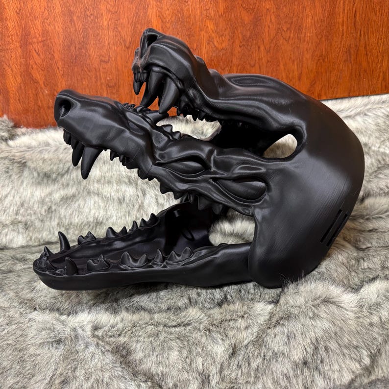 May include: A black, stylized mask in the shape of a snarling canine skull. The mask features detailed teeth, a textured surface, and a menacing expression. The mask is displayed on a faux fur surface.