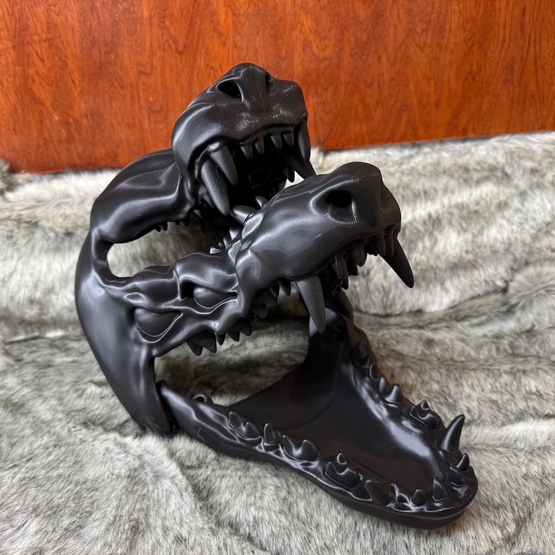 May include: A black sculpture of three stylized dragon skulls with sharp teeth. The sculpture is made of a glossy material and is set against a textured, gray background.