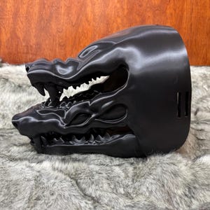 May include: A black, 3D-printed mask shaped like a snarling beast's head. The mask features sharp teeth and detailed facial features. The mask is resting on a gray, furry surface.