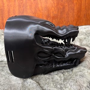 May include: A black, 3D-printed mask in the shape of a snarling beast's head. The mask features sharp teeth, detailed facial features, and three vertical slits on the side. The mask is made of a shiny, smooth material.
