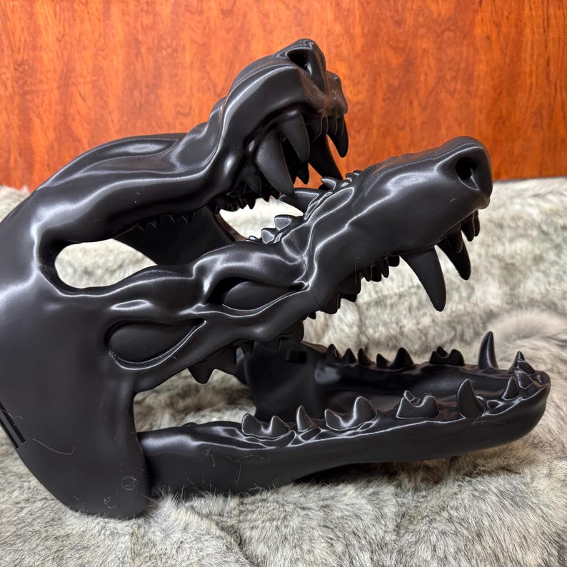May include: A black, stylized sculpture of a canine skull, featuring detailed teeth and a textured surface. The sculpture is set against a backdrop of faux fur and a wooden panel.