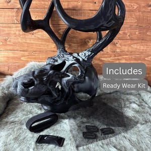 May include: A black deer skull mask with large antlers, part of a ready-to-wear kit. The mask has a detailed, textured design with white accents. Includes a black strap, mesh, and buckles.