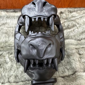 May include: A black, 3D-printed sculpture of a large animal skull, featuring prominent teeth and detailed texture. The skull is mounted on a black stand, set against a backdrop of faux fur.