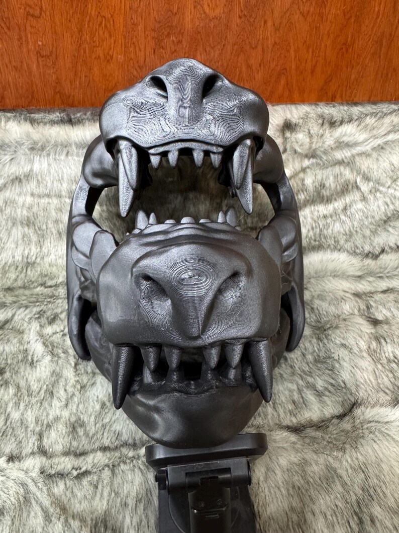 May include: A black, 3D-printed sculpture of a large animal skull, featuring prominent teeth and detailed texture. The skull is mounted on a black stand, set against a backdrop of faux fur.