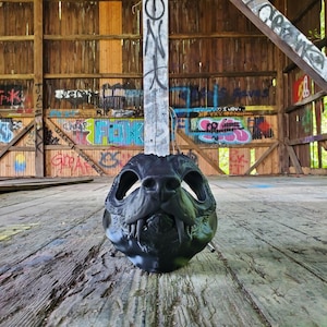 May include: A black, stylized mask resembling a canine skull, featuring detailed teeth and eye sockets. The mask is set against a backdrop of a weathered wooden structure with graffiti art, creating a contrast between the mask's dark form and the rustic environment.