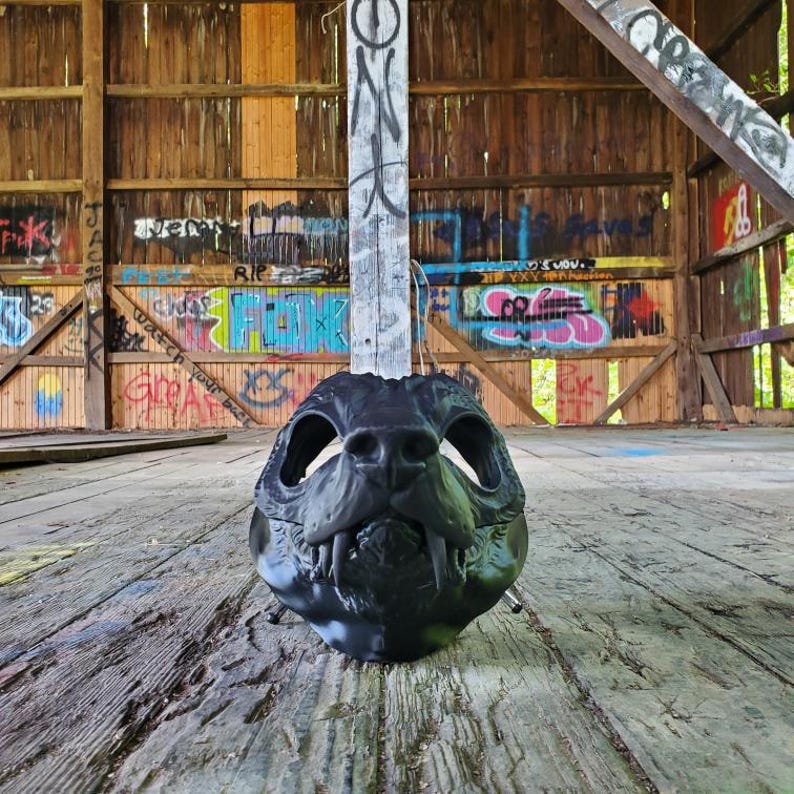 May include: A black, stylized mask resembling a canine skull, featuring detailed teeth and eye sockets. The mask is set against a backdrop of a weathered wooden structure with graffiti art, creating a contrast between the mask's dark form and the rustic environment.