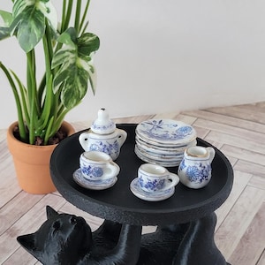 Puede incluir: Una figura de gato negro que sostiene una bandeja redonda en su espalda, coronada con un juego de té de porcelana en miniatura con patrones florales azules y bordes dorados. El juego incluye una tetera, dos tazas con platillos y una lechera.