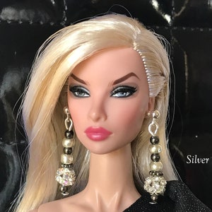 May include: A doll with blonde hair wearing silver earrings. The earrings have a cluster of clear crystals at the bottom and a mix of silver and pearl-colored beads. The text "Silver" is visible.