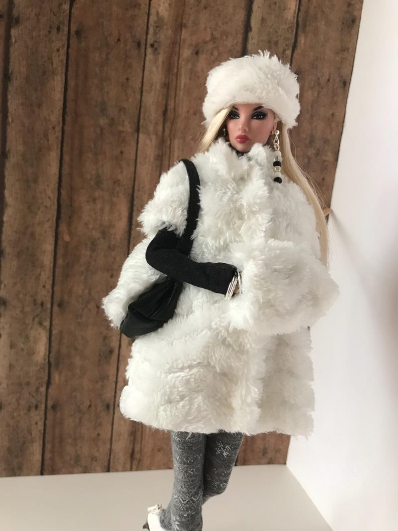 May include: A white faux fur coat with a white faux fur hat and a black handbag. The doll is wearing grey tights and white shoes.