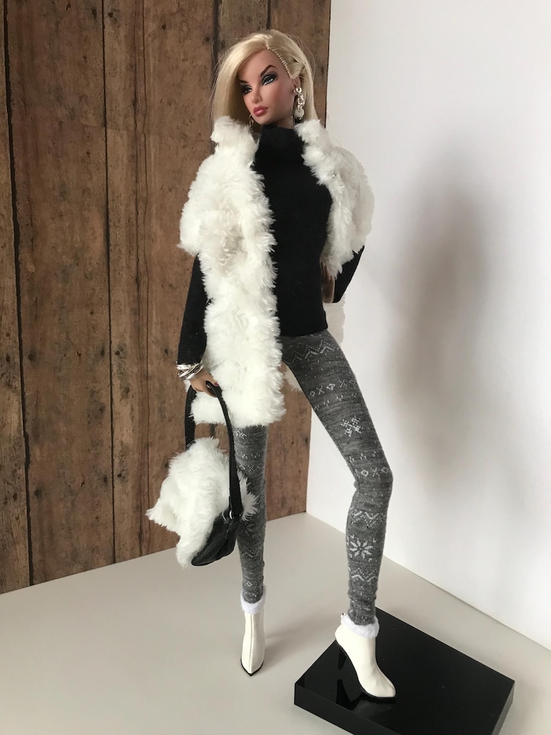 May include: A fashion doll wearing a white faux fur vest, a black turtleneck sweater, gray leggings with a snowflake pattern, and white boots. The doll is holding a white faux fur handbag with a black strap.
